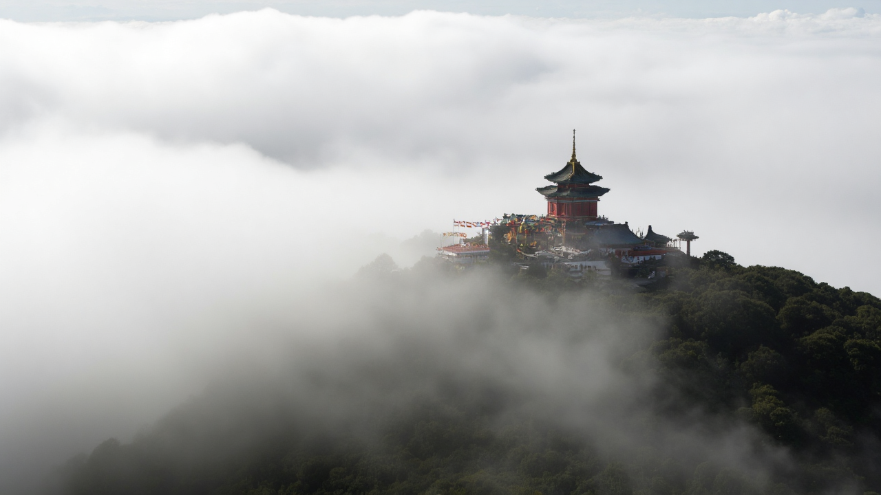 Ancient temple structures at Mount Emei’s Golden Summit emerge from swirling clouds, adorned with colorful Tibetan prayer flags Golden Summit temple complex atop Mount Emei shrouded in clouds with Buddhist prayer flags fluttering