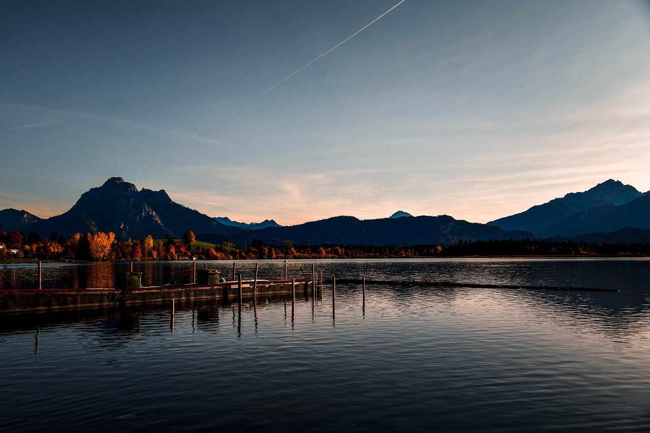 Hopfensee lake near Füssen — mirror-still waters reflecting snow-capped Alps and a solitary wooden rowboat — a serene, reflective finale among the 15 must-visit places in Germany. Hopfensee lake with alpine backdrop, rowboat, and clear blue water