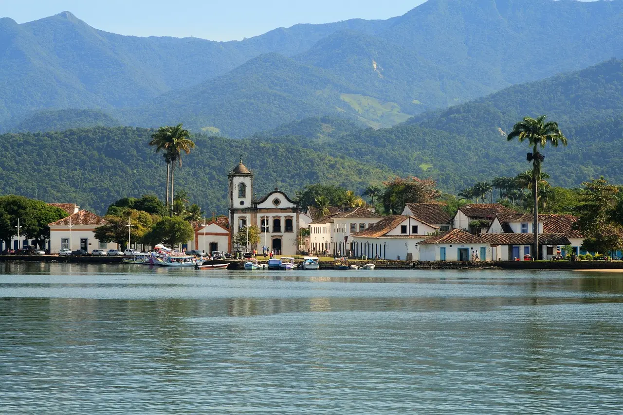 Historic colonial waterfront in Paraty, Rio de Janeiro — pastel buildings reflected in calm harbor waters, classic schooners docked beneath tropical palms and granite cliffs Colonial waterfront with colorful buildings and sailing boats in Paraty
